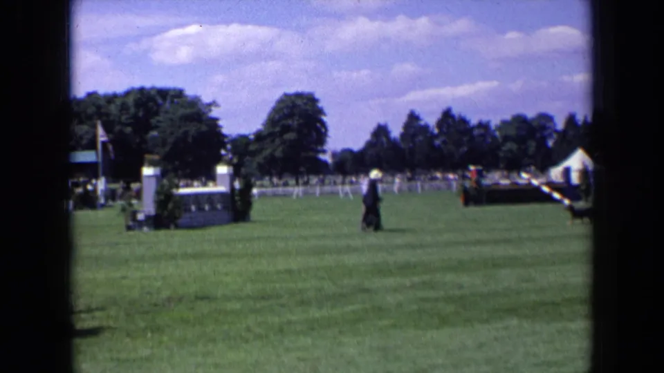 1963 England frame with a suited man on a broad grassy show ground, with equestrian fencing, trees, and a white tent visible beyond him.