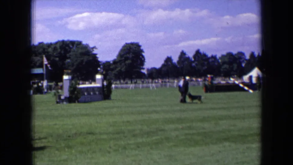 1963 England home-movie frame showing a black dog approaching a suited man on the lawn, with event fencing, trees, and a tent behind them.