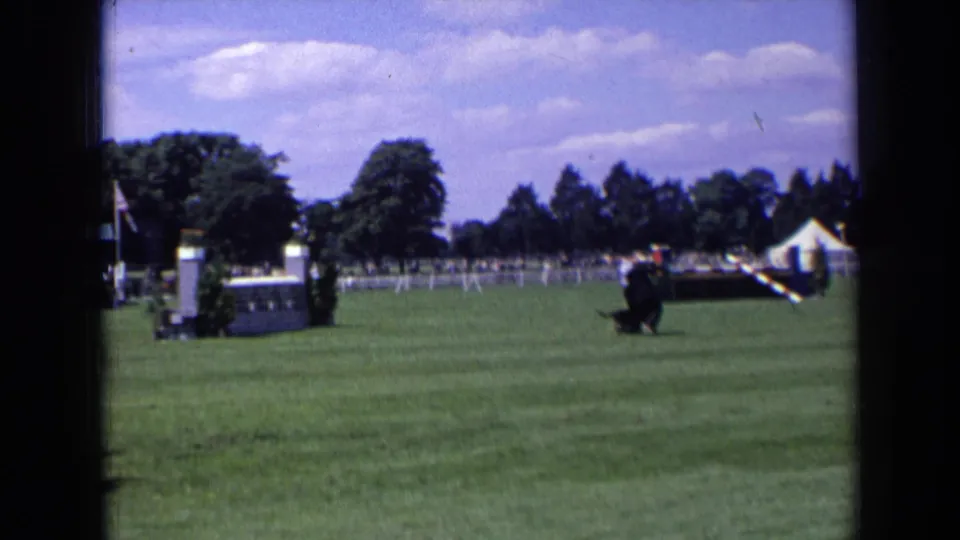 1963 England frame showing a suited man and black dog together on the show-ground lawn, with white fencing, trees, and tented event structures behind.