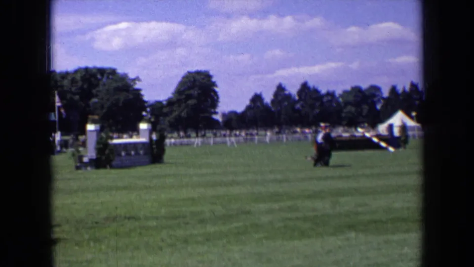 1963 England home-movie frame showing a rider on horseback near a white jump during an outdoor equestrian event.