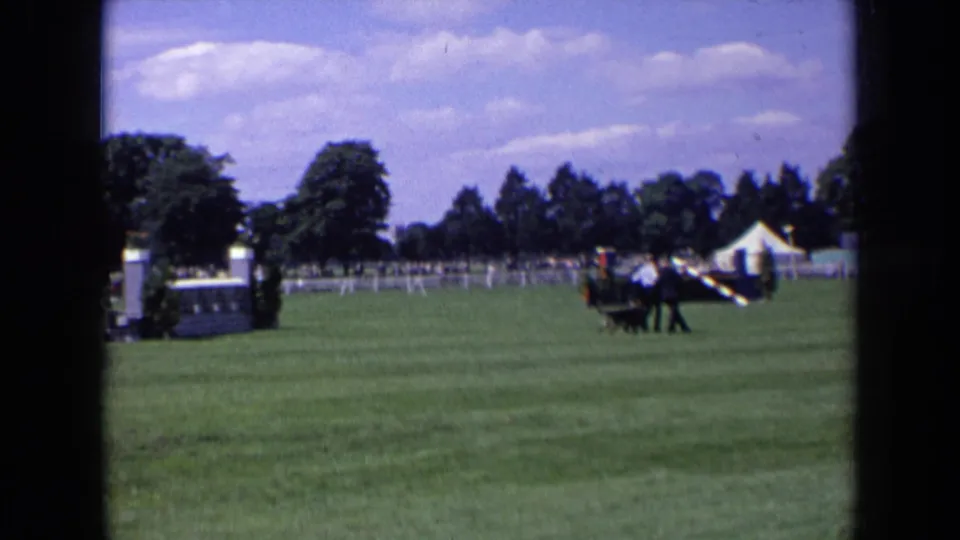 1963 England equestrian event frame with a horse and rider moving past a white jump and distant spectators.