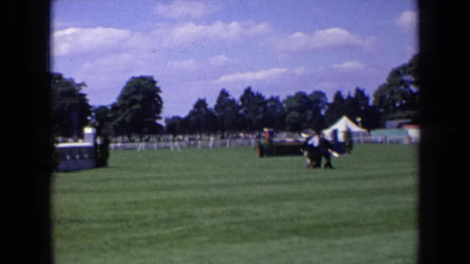 1963 England home-movie frame of a horse and rider near a low white jump in an outdoor competition field.