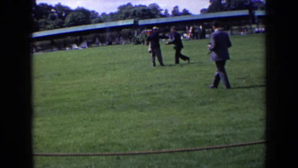 1963 England event frame showing suited men walking across the grass, one appearing to carry a camera, near spectators.