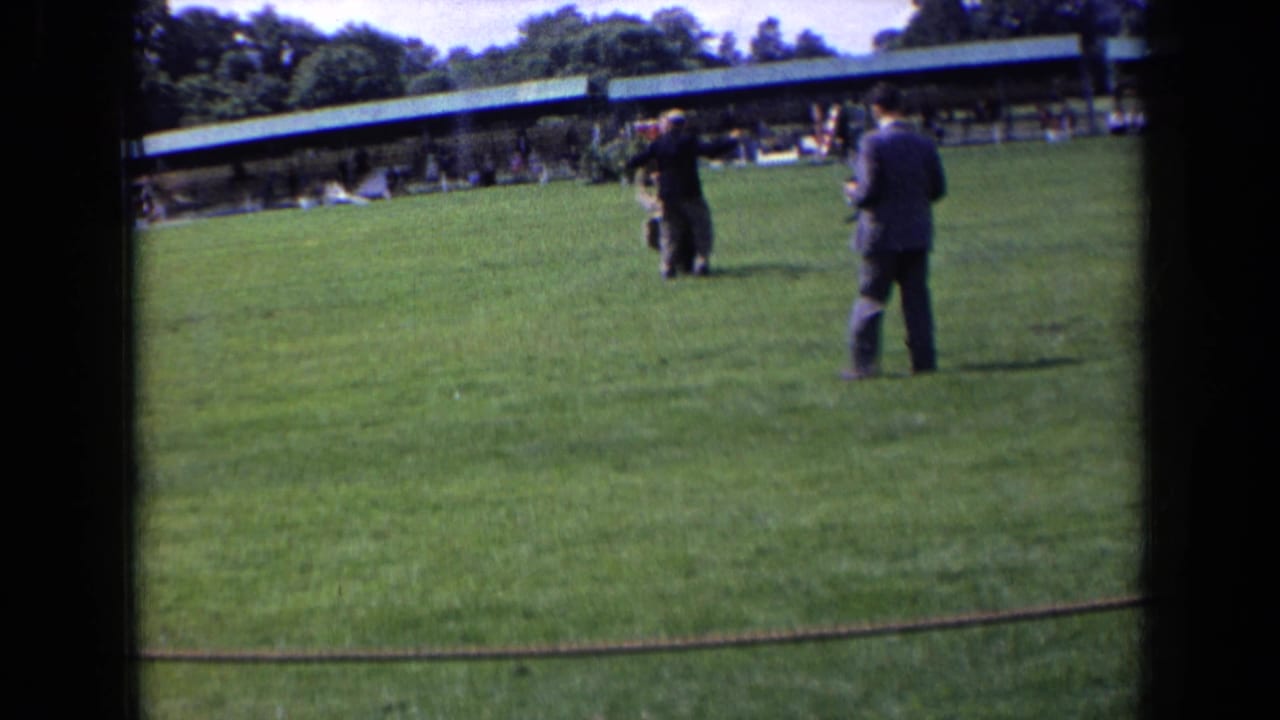 1963 England home-movie frame showing men in dark suits and coats on a grassy lawn with spectators under a pavilion.