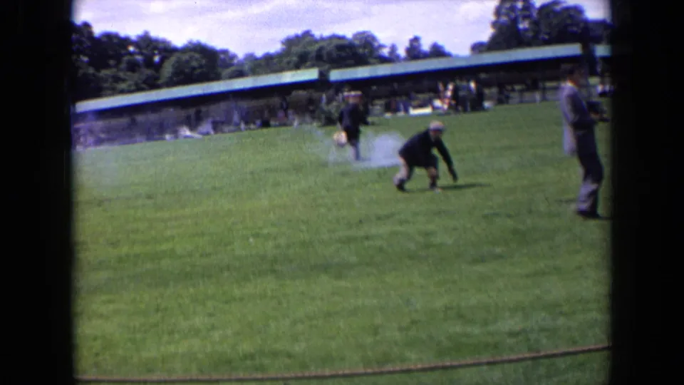 1963 England home-movie frame showing a man crouching or falling on the lawn as starter gun smoke drifts behind him.