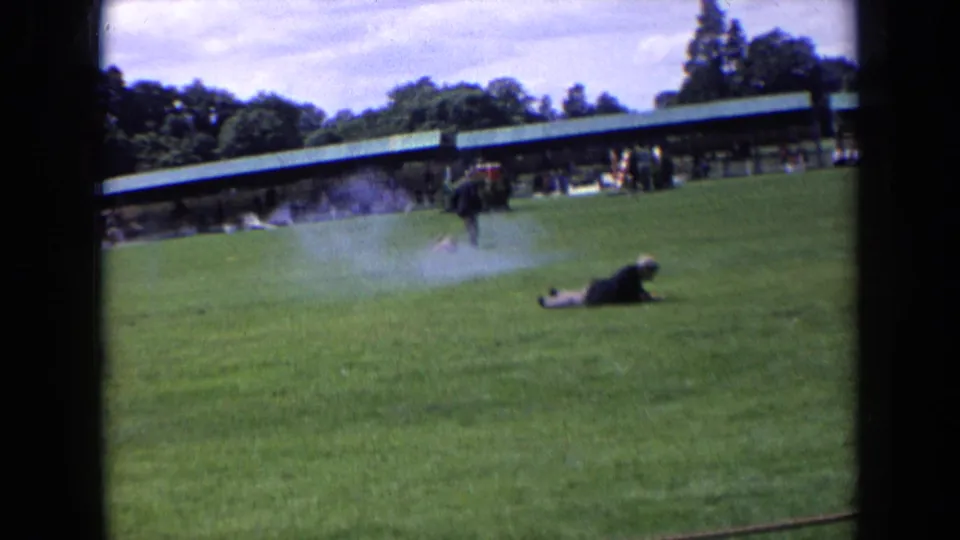1963 England frame showing a man lying on a grassy lawn with smoke drifting and other figures near the pavilion in the background.