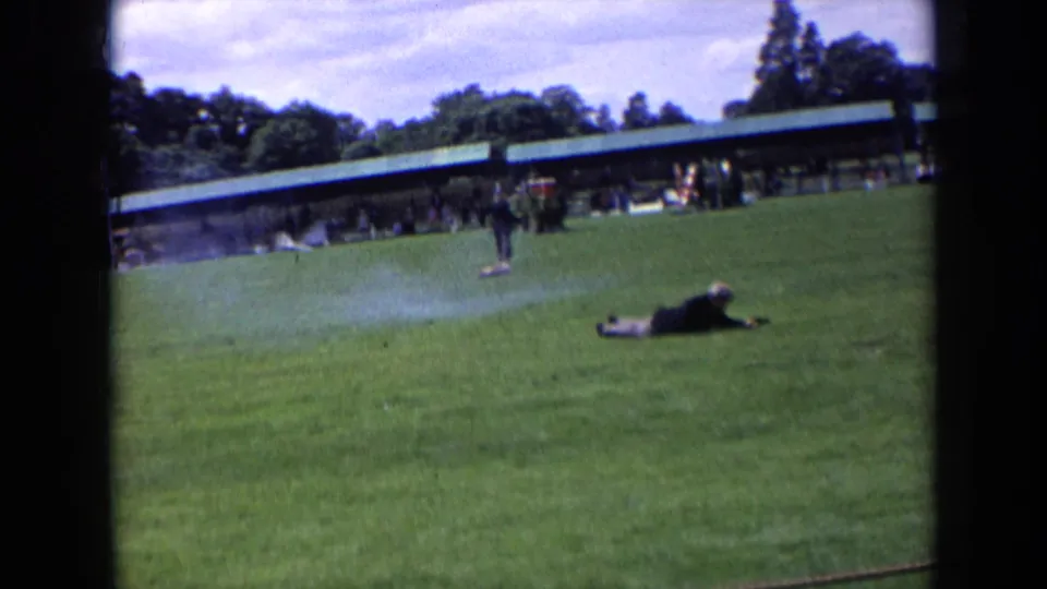 1963 England 8mm frame showing a dog near a person lying on a grass sports field, with spectators under a long green-roof shelter.