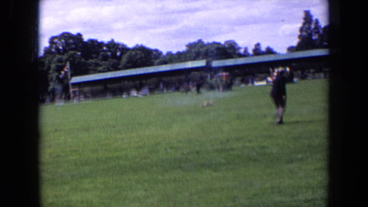 A rider crosses a grassy show-jumping field in front of a long pavilion.
