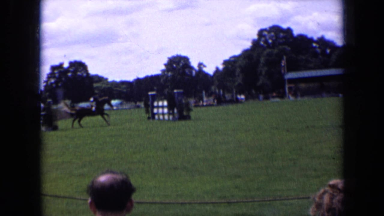 1963 England home-movie frame with a horse and rider near a jump, a rope barrier, seated spectators and trees behind the course.