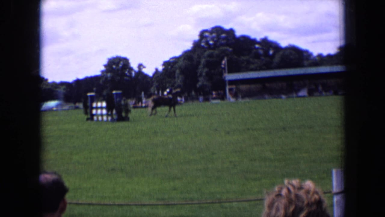 1963 England frame showing a horse and rider clearing or passing a jump, with spectators, trees, pavilion roof and rope barrier visible.