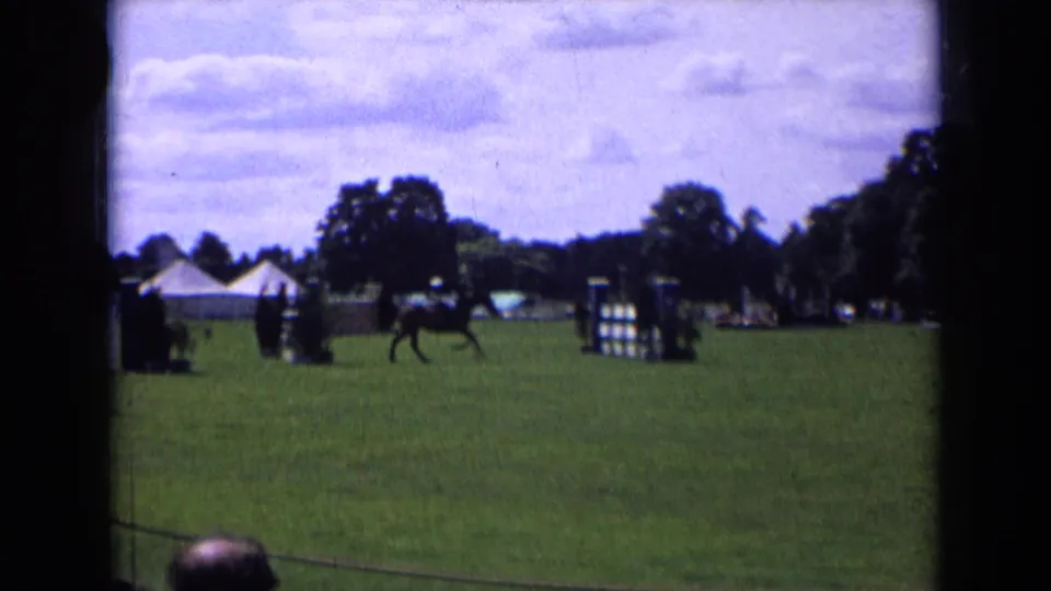 In 1963 England, a horse and rider cross a grass equestrian arena with striped jumps, trees, tents, and distant spectators visible.
