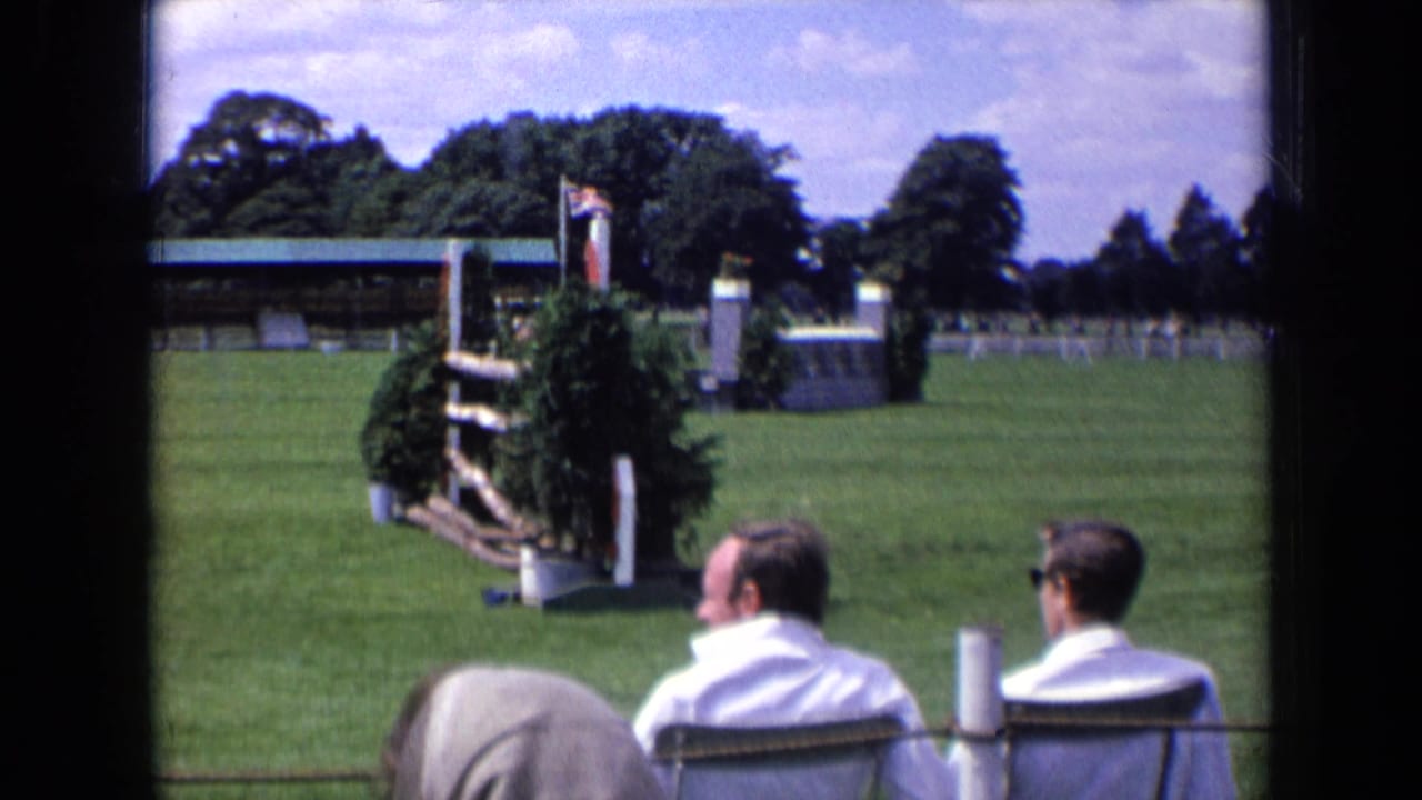 Spectators in white shirts sit behind a rope barrier as a decorated hedge jump and pavilion stand on the show-jumping field.