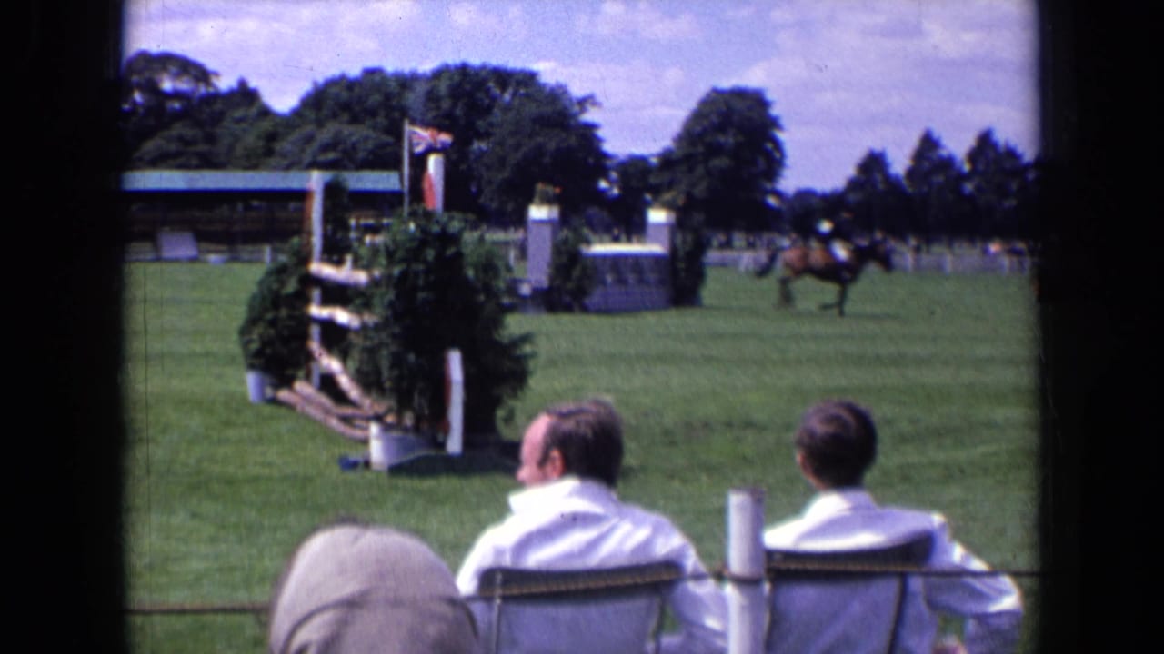 At the 1963 England equestrian event, foreground spectators watch as the horse and rider pass beyond a decorated jump on the grass arena.