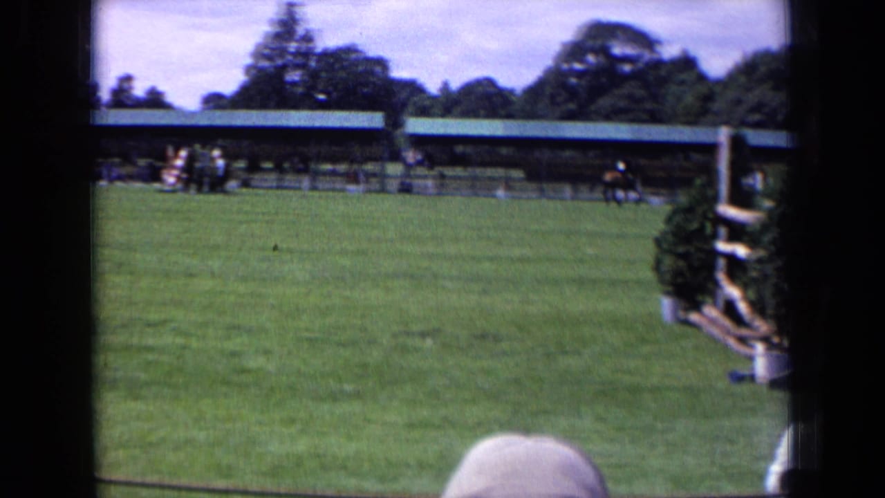 Home-movie frame of a horse and rider approaching or clearing a fence at an English equestrian ground, with pavilion and spectators visible.