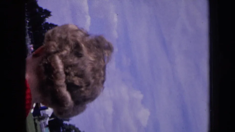 Sideways 8mm frame from the same 1963 England clip, showing a nearby woman in close foreground as the camera shifts away from the field.