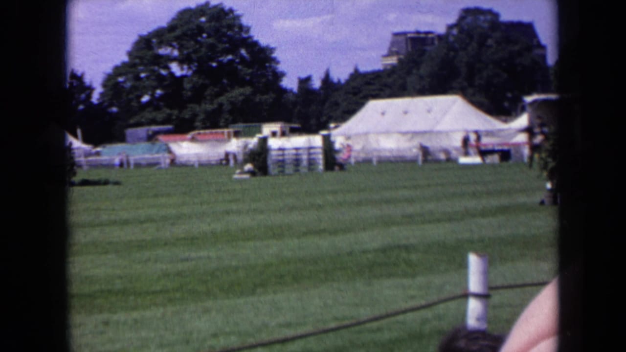 Wide grass arena with event tents, fence sections, and a horse near the right edge.