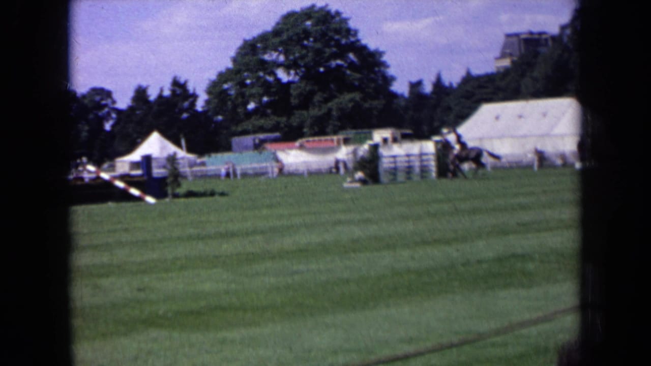 1963 England home-movie frame of an equestrian competition course with striped rails, white tents, and a mounted rider moving past the jumps.