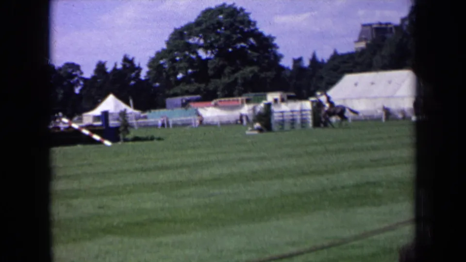 1963 England home-movie frame of an equestrian competition course with striped rails, white tents, and a mounted rider moving past the jumps.