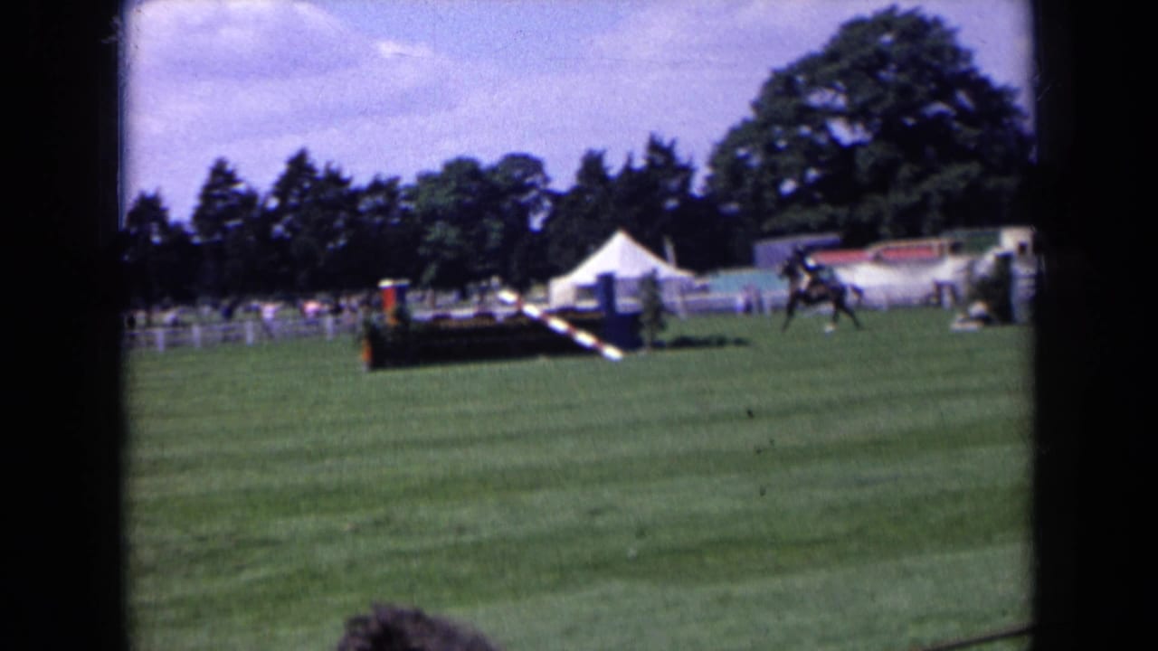 A distant 1963 England equestrian competition view shows the horse and rider crossing the field beyond a striped obstacle and white tents.