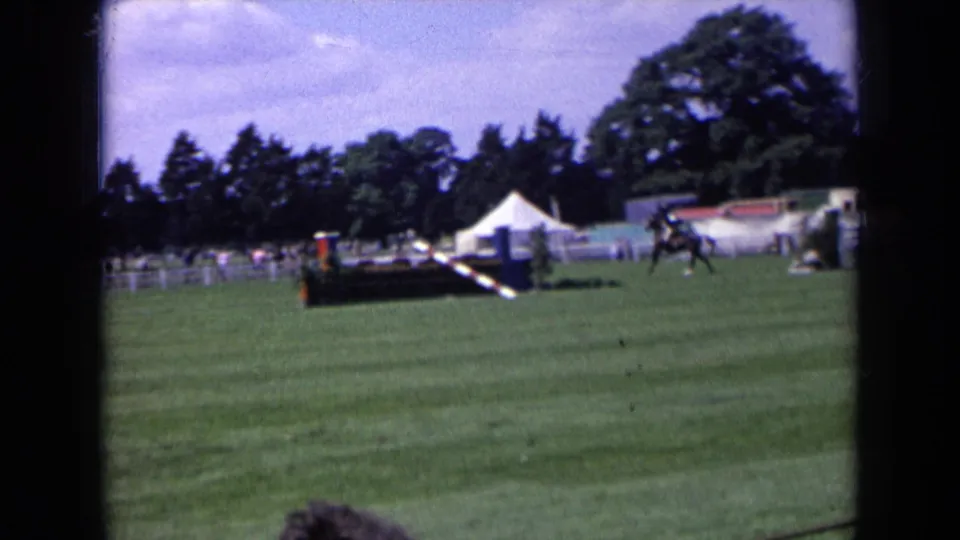 A distant 1963 England equestrian competition view shows the horse and rider crossing the field beyond a striped obstacle and white tents.