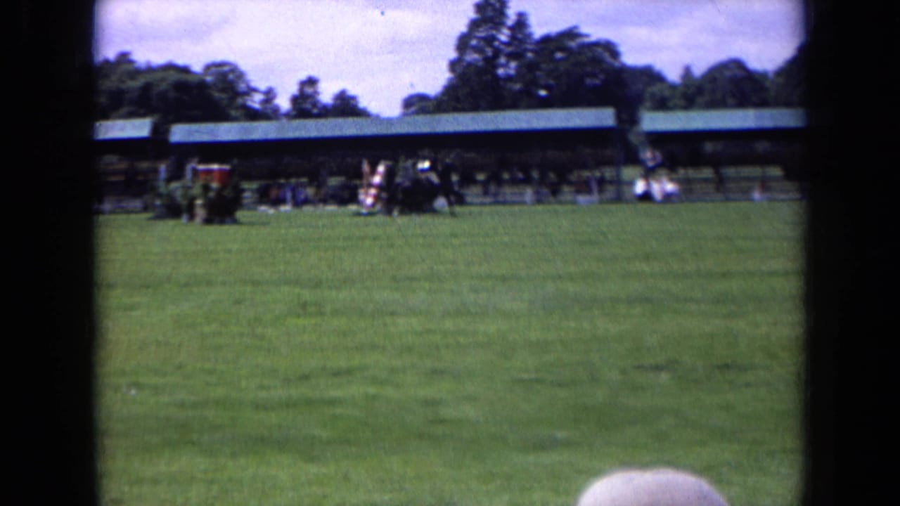 Distant 8mm frame of a horse and rider at an outdoor riding show in England, with jump equipment and spectators in the background.