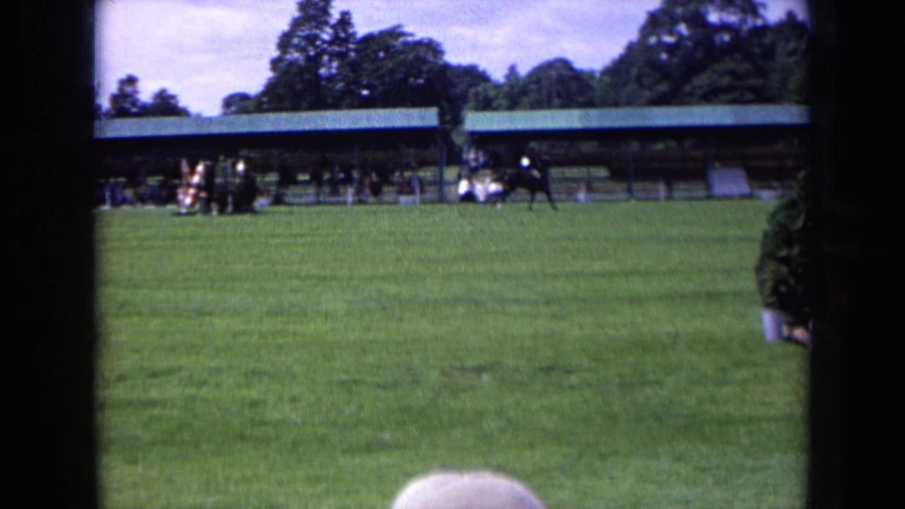 The horse and rider continue across the 1963 England riding field, framed by grass, jump equipment, a covered stand, and distant spectators.