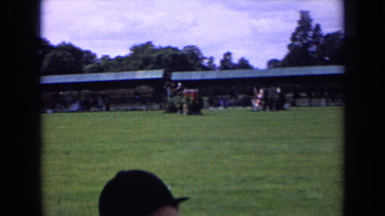 Wide 8mm view of an English equestrian field with distant people near covered buildings and a spectator partly blocking the foreground.