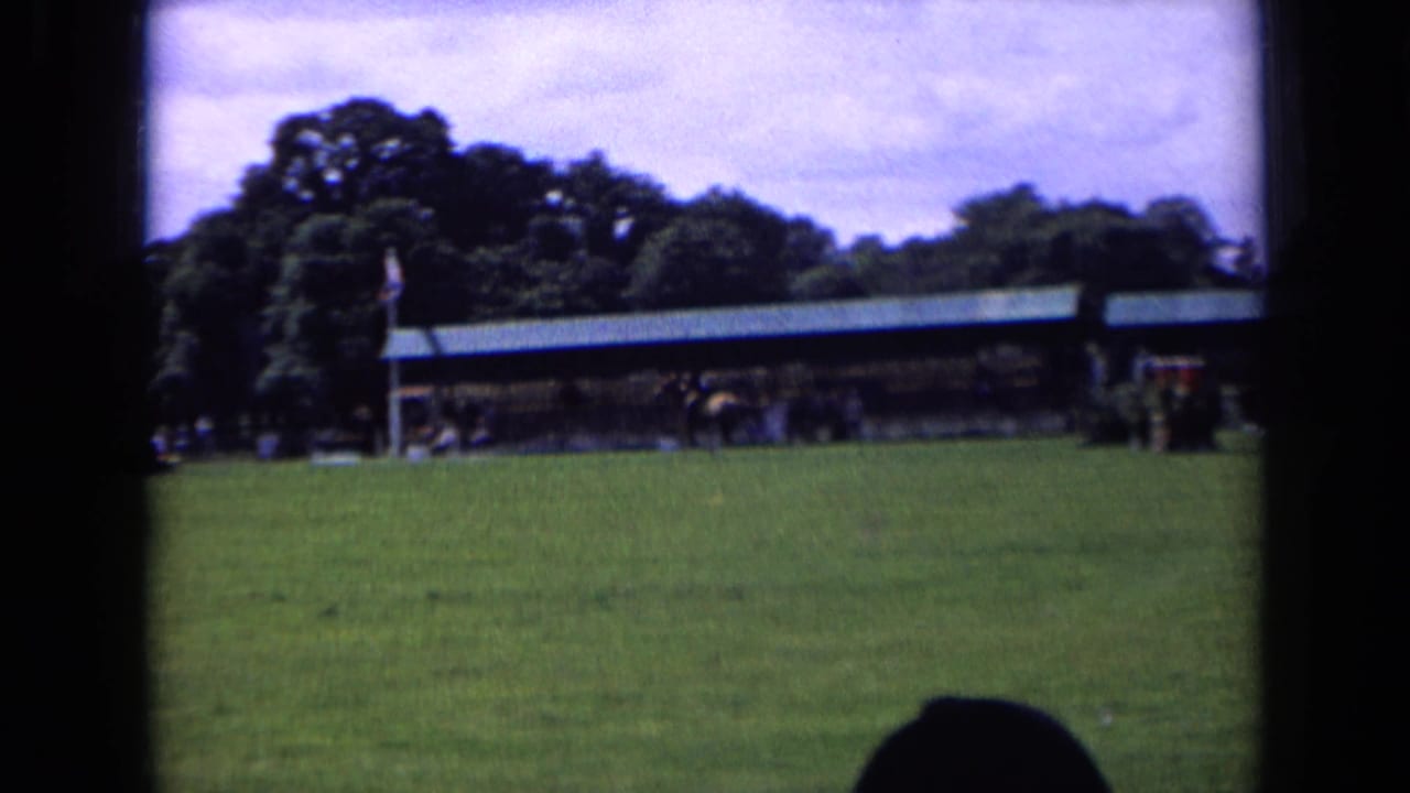 Handheld 8mm frame from an England horse show, looking across grass toward covered event buildings, trees, and distant spectators.