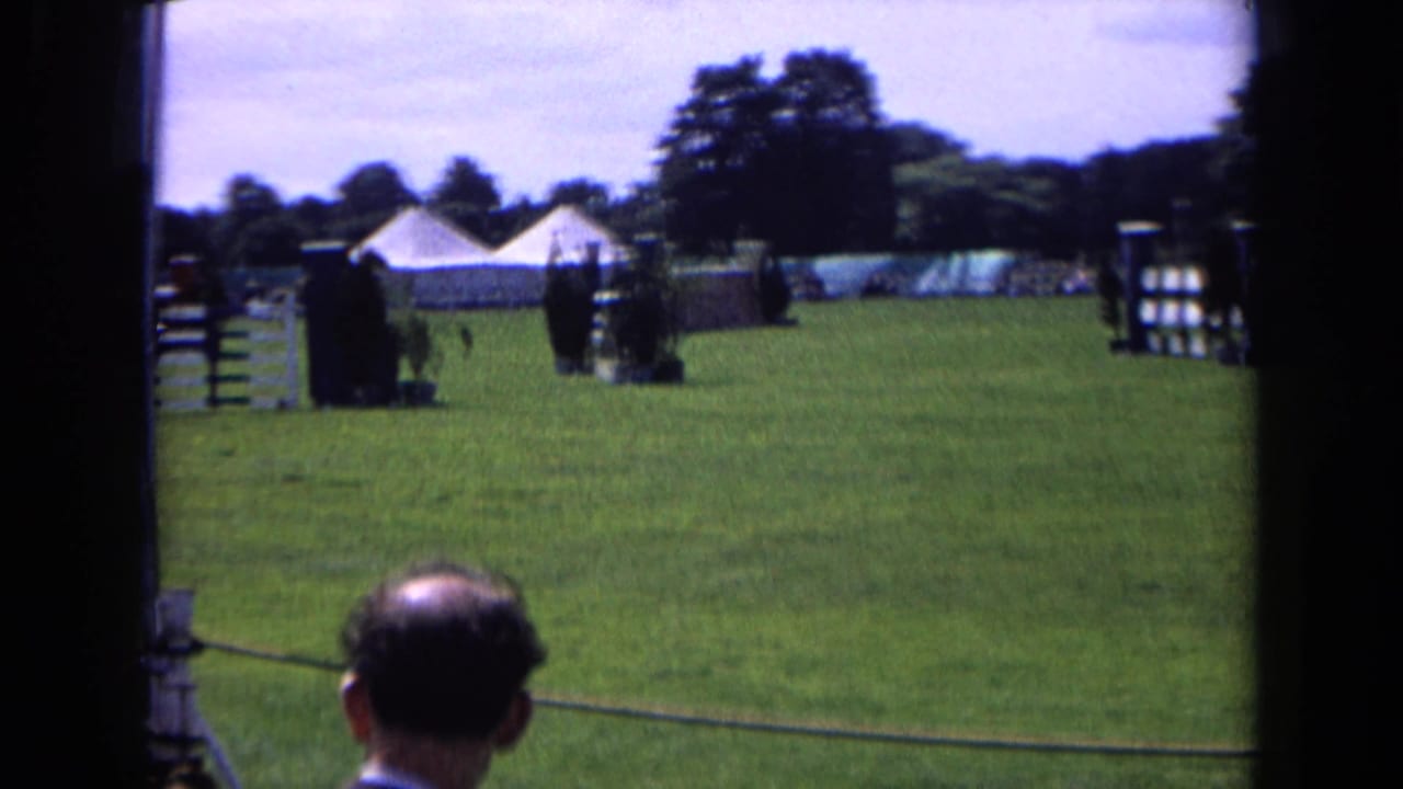 1963 England equestrian field with jump standards, white tents, rope barrier, trees, and spectators viewed from near the sideline.