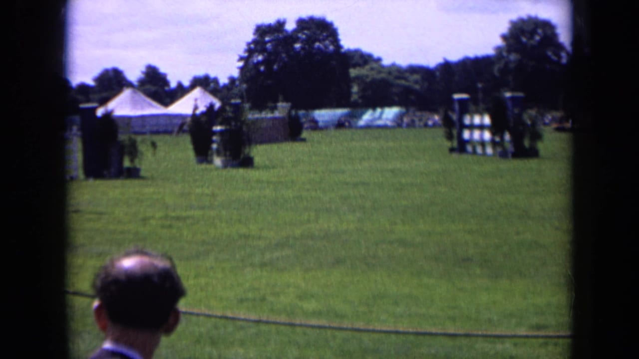 1963 England 8mm frame showing an equestrian course, spectators at the rope barrier, white tents, trees, and jump obstacles on a sunny field.