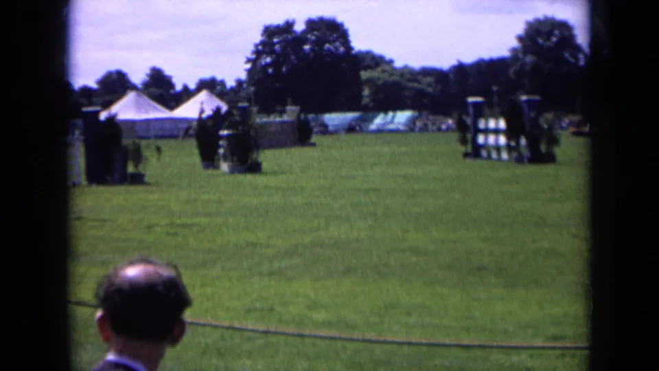 1963 England 8mm frame showing an equestrian course, spectators at the rope barrier, white tents, trees, and jump obstacles on a sunny field.
