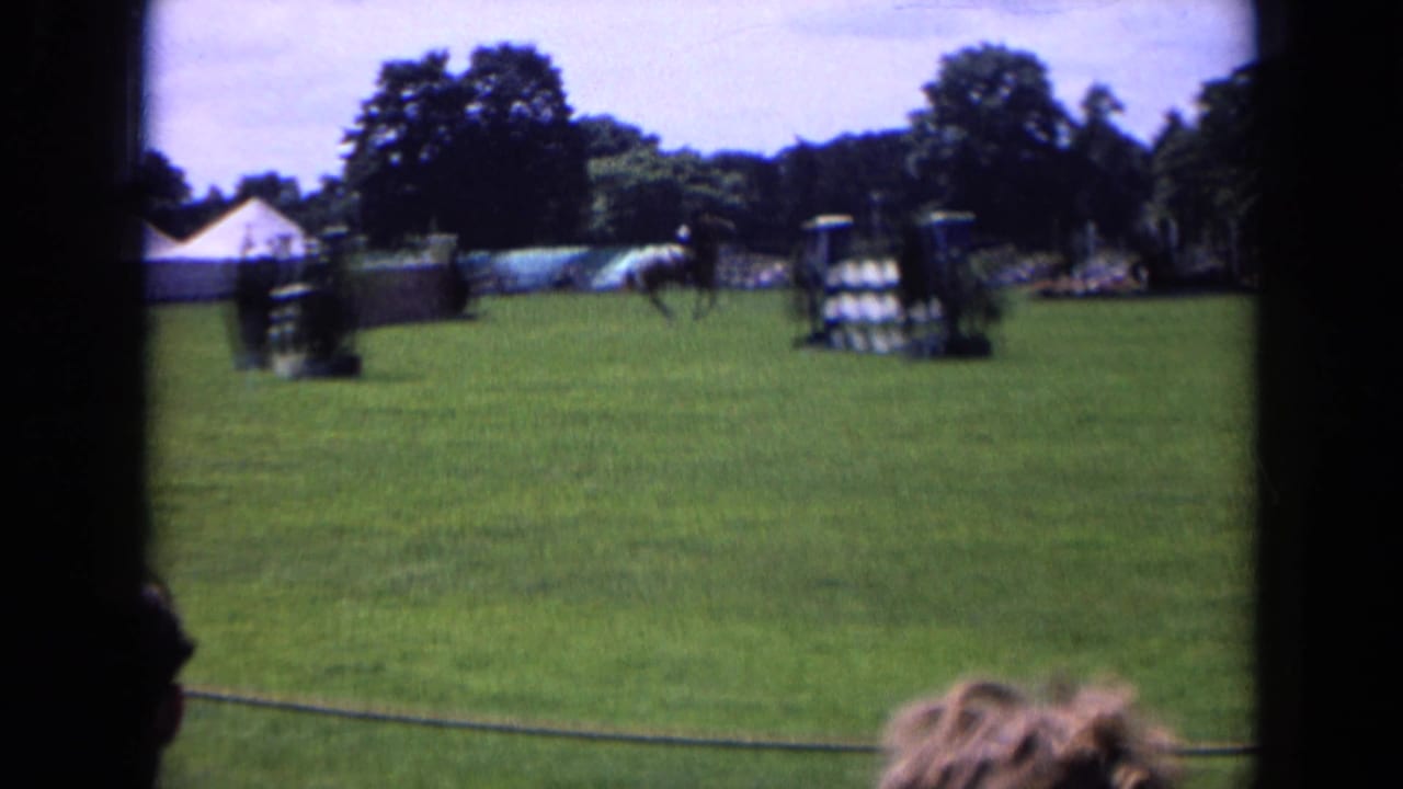 Rider and horse move across a grassy equestrian course with tents and jumps in the background.