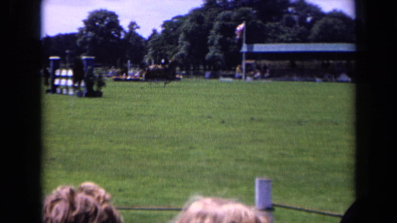 Wide 8mm view of an English equestrian field in 1963, with jumps, a covered viewing area, spectators, trees, and foreground heads.