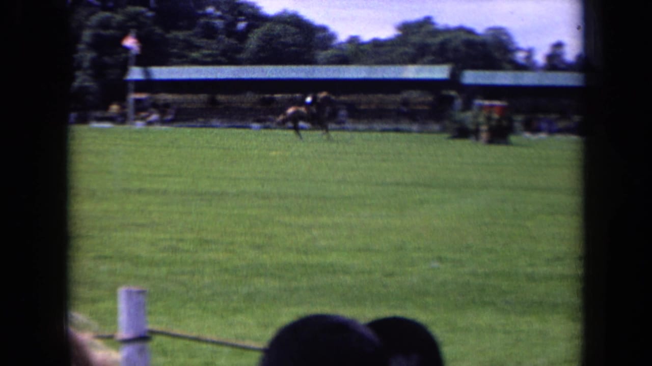 1963 England home-movie frame showing a horse and rider crossing the far side of an equestrian field with spectators in the foreground.