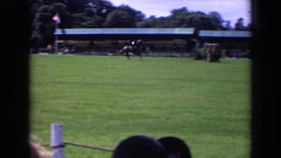 1963 England home-movie frame showing a horse and rider crossing the far side of an equestrian field with spectators in the foreground.