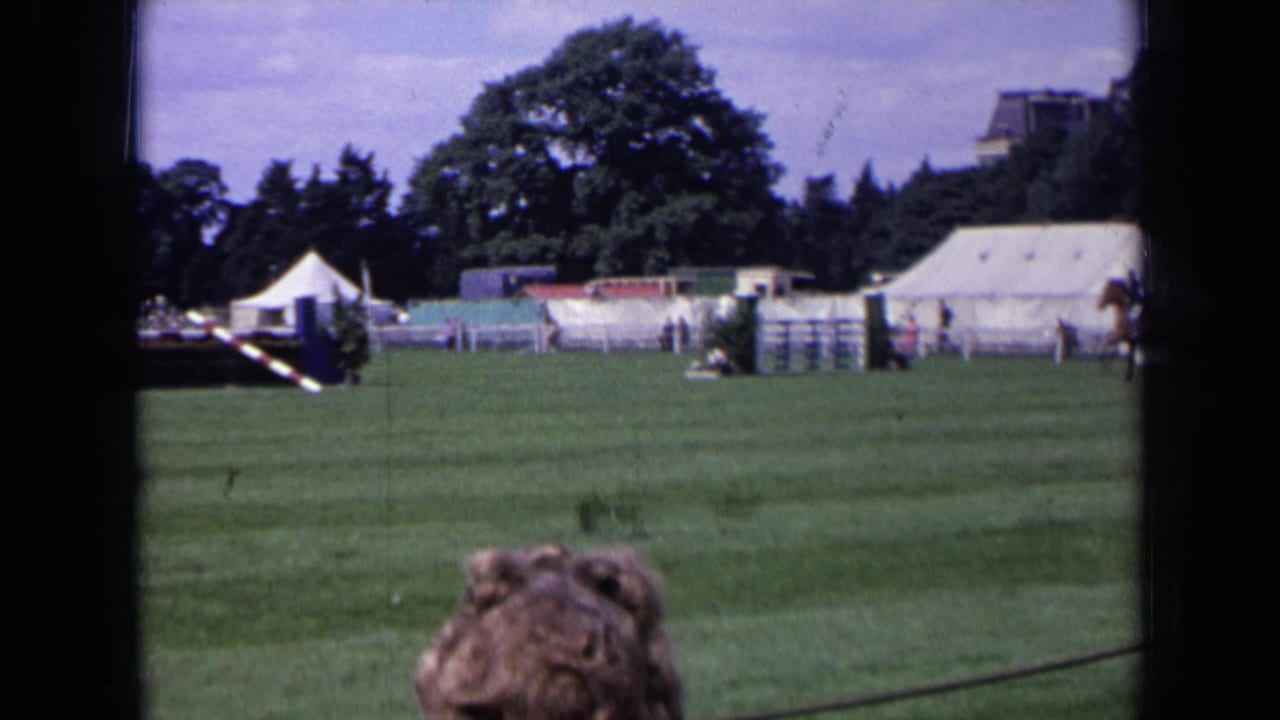1963 England home-movie frame with a wide grass-field view, white tents, trees, and foreground spectators at an outdoor riding event.