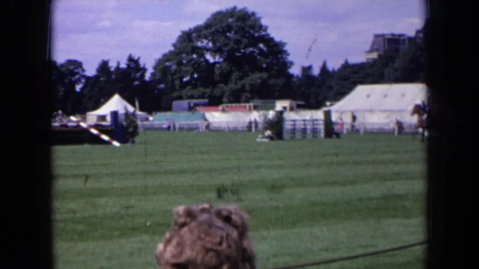1963 England home-movie frame with a wide grass-field view, white tents, trees, and foreground spectators at an outdoor riding event.