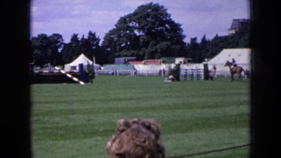 1963 England 8mm view of an equestrian show field with jump rails, white tents, trees, and a foreground spectator.
