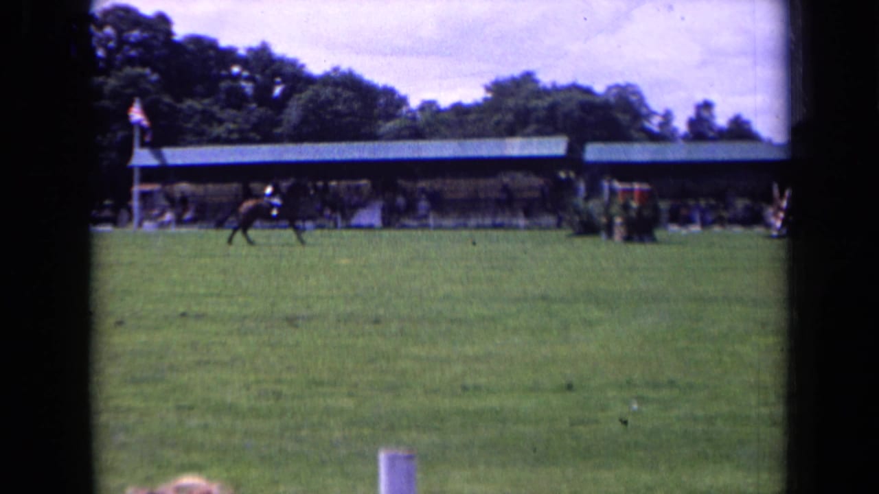 A horse and rider gallop across the 1963 England showground, with a long covered structure, trees, and spectators in the background.