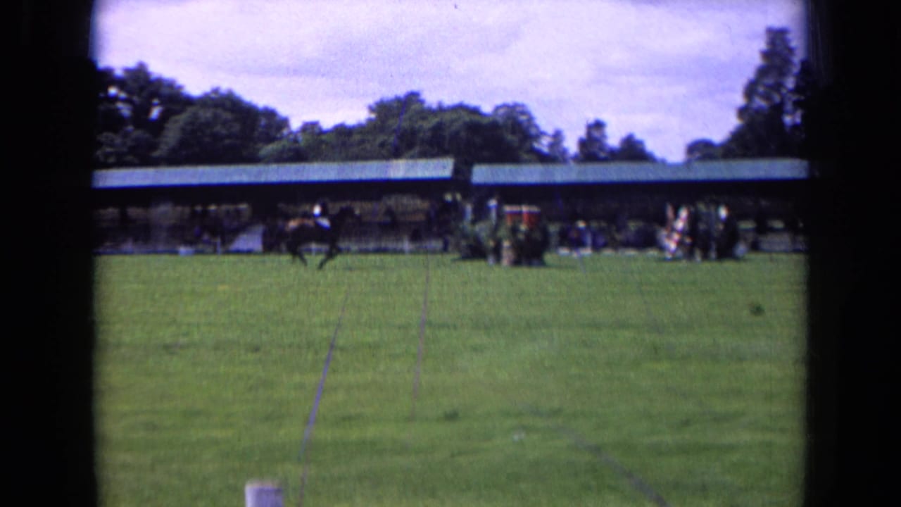1963 England 8mm frame showing a grass equestrian field with spectators gathered near a long covered structure.