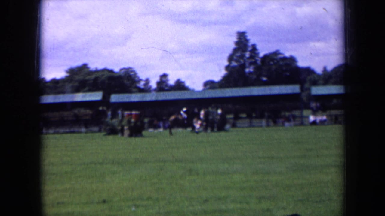 A horse and rider cross the grass show arena in 1963 England, with spectators and a covered stand behind them.
