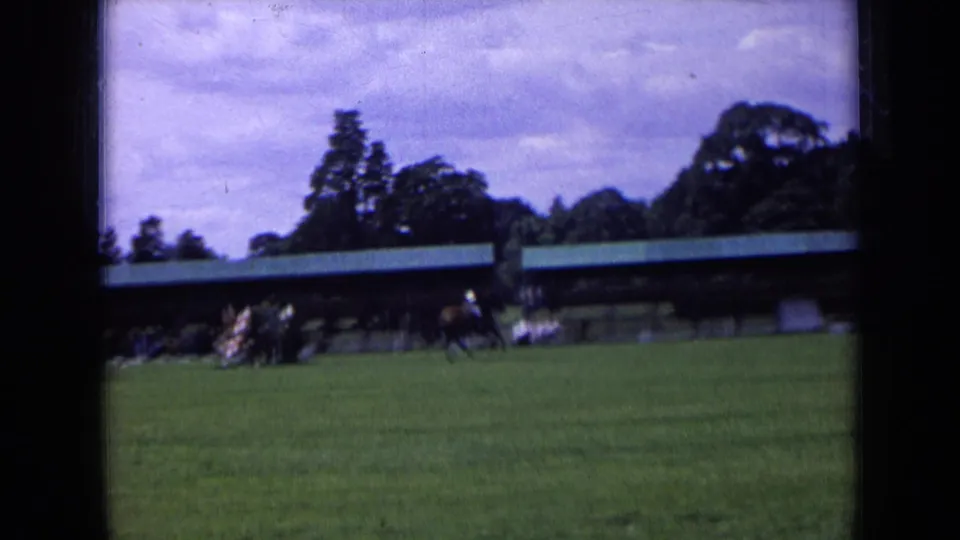 Soft 8mm frame of a distant equestrian scene on a grass field in England, with spectators near a covered viewing area.