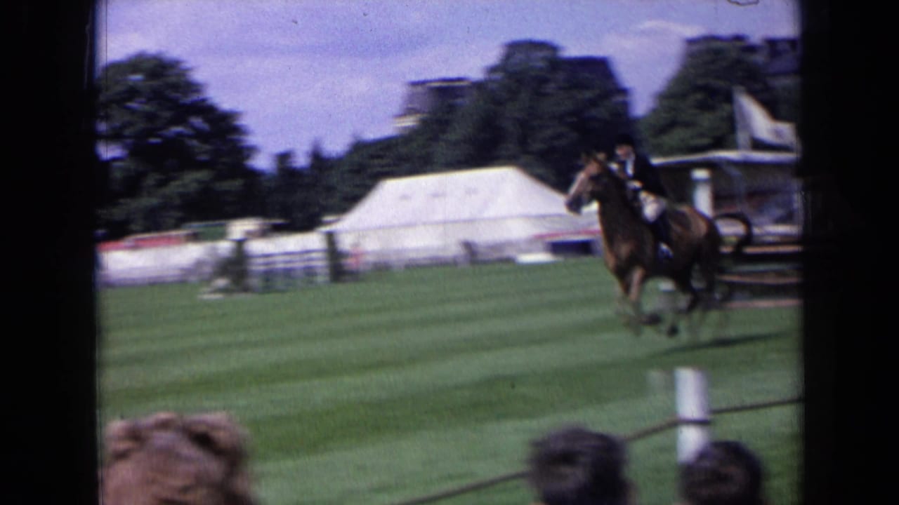 In 1963 England, a mounted rider guides a brown horse across a grass show-jumping field as tents and spectators frame the arena.