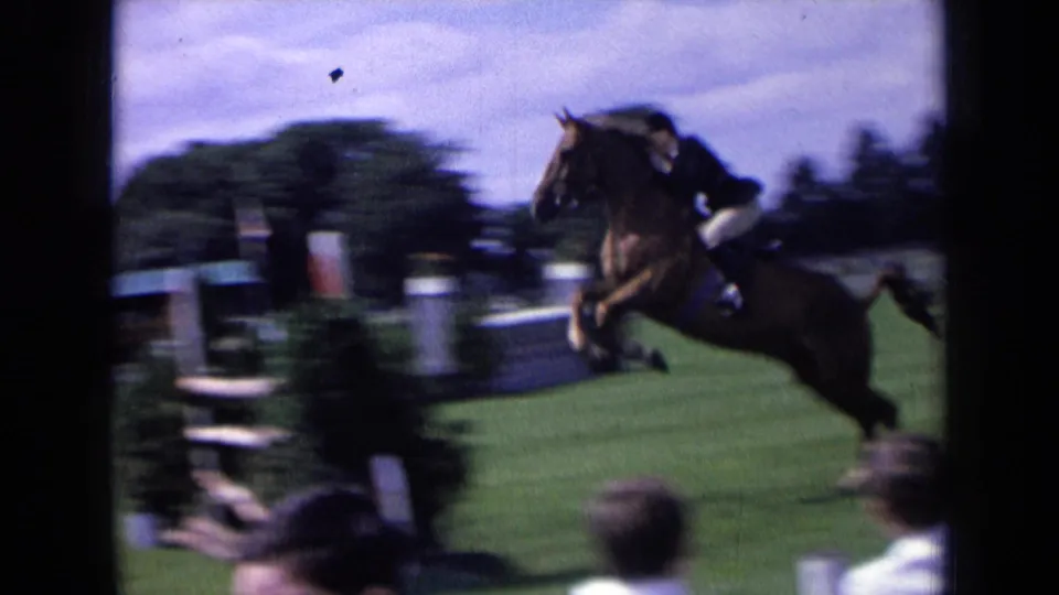 A brown horse with a rider in show attire clears an obstacle on a 1963 England show-jumping field with spectators nearby.