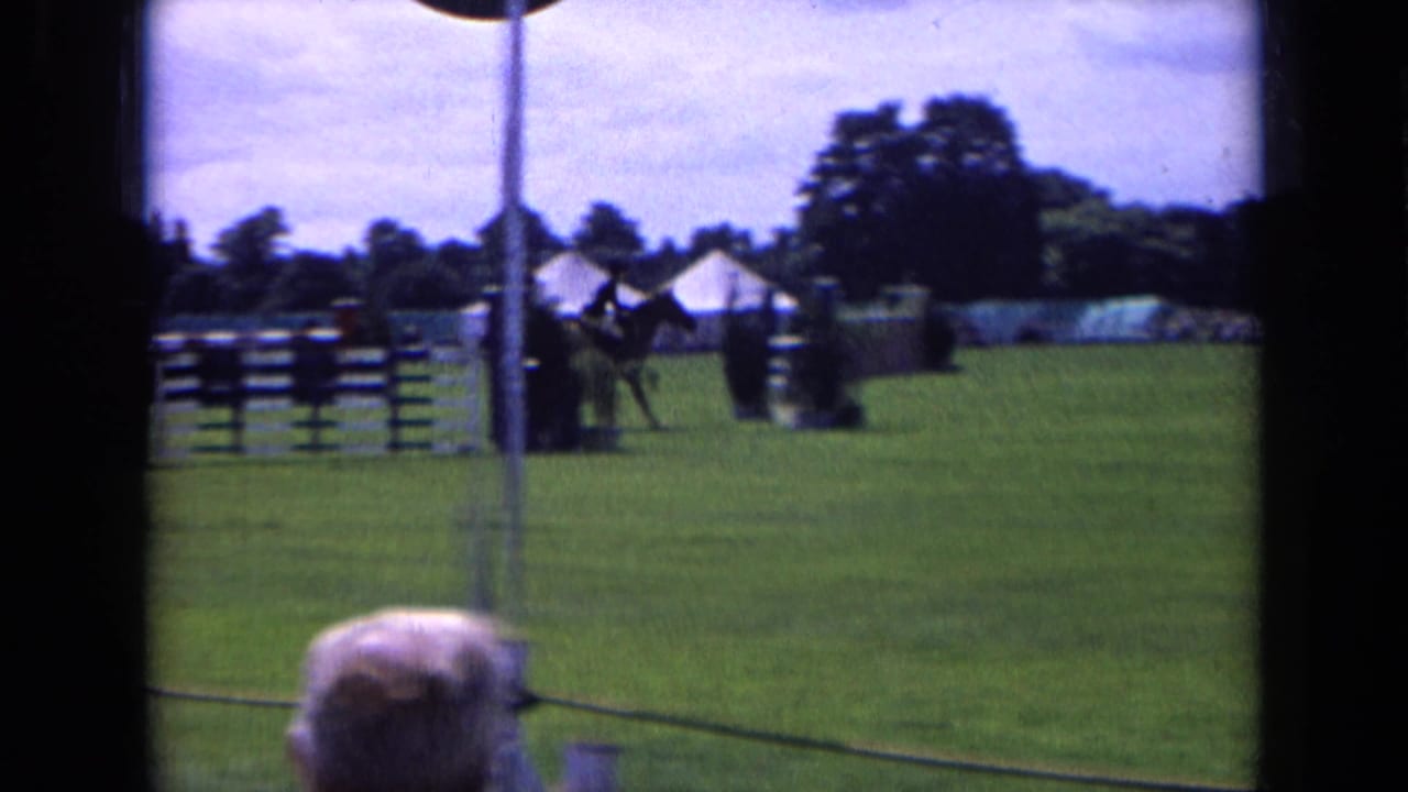 1963 England 8mm frame of a horse and rider in a grassy show ring, with jump fences, tents, trees, and spectators behind a rope barrier.