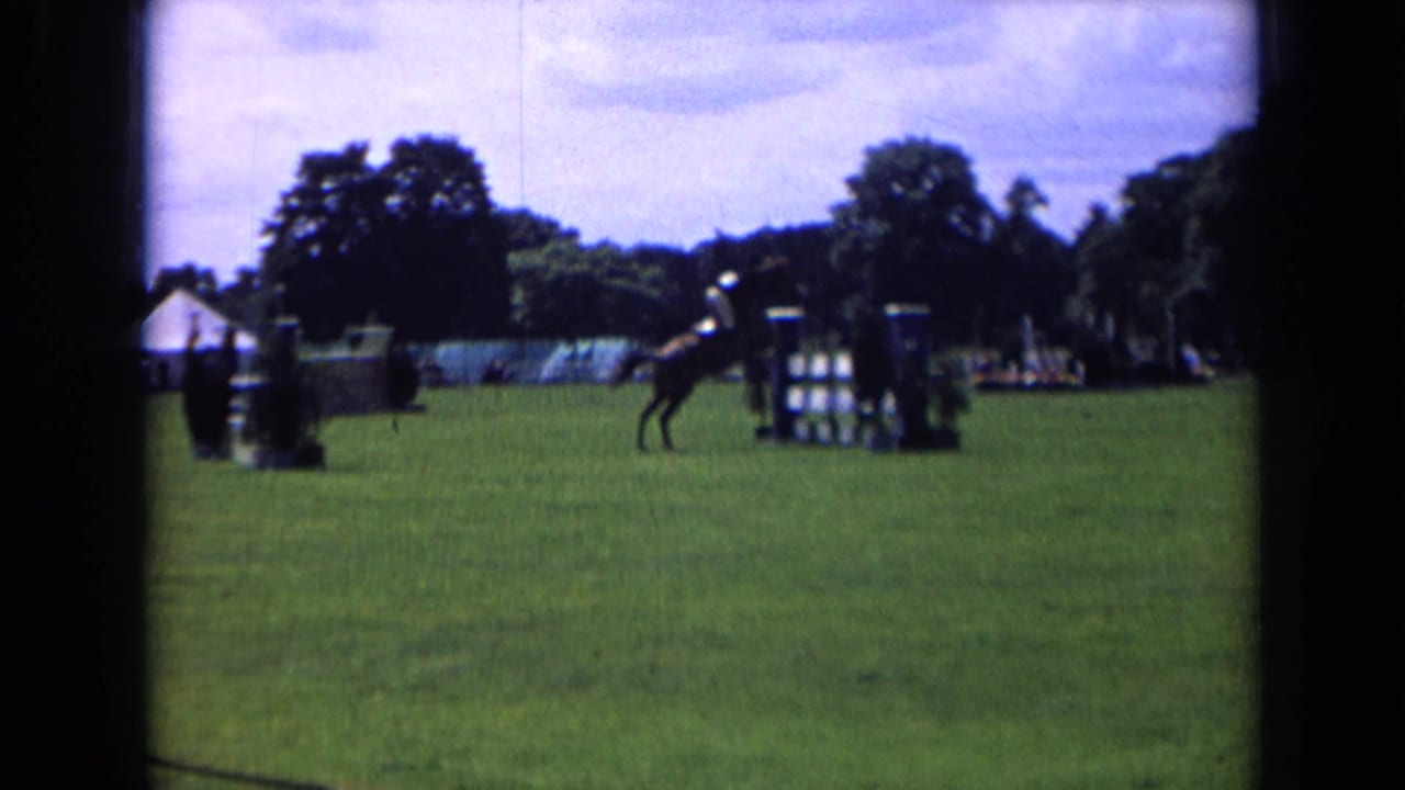 Mounted rider approaches a blue-and-white jump fence on a grassy field with tents in the distance.