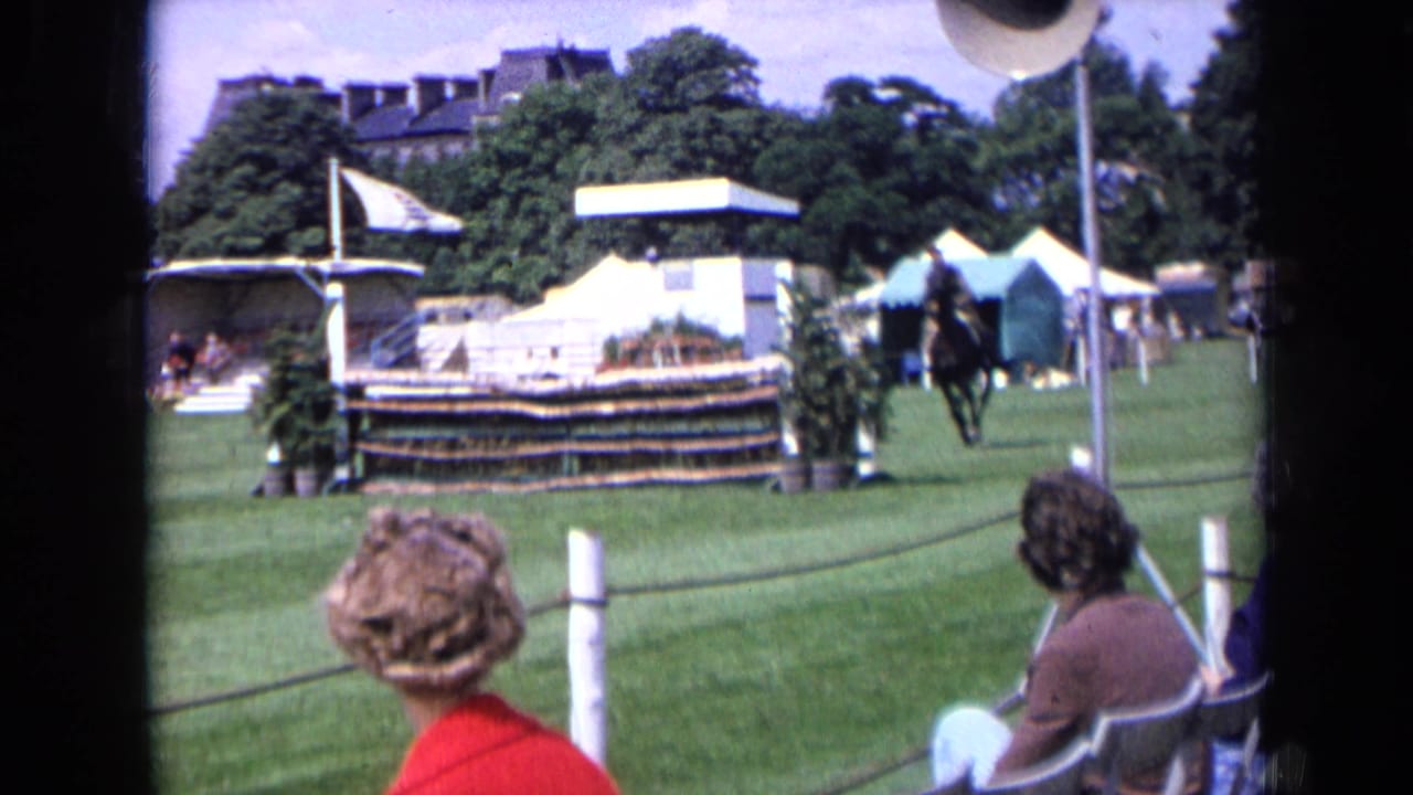 1963 England 8mm frame from the spectator side of a horse show, with seated viewers, rope barrier, white tents, and a rider near the jump area.