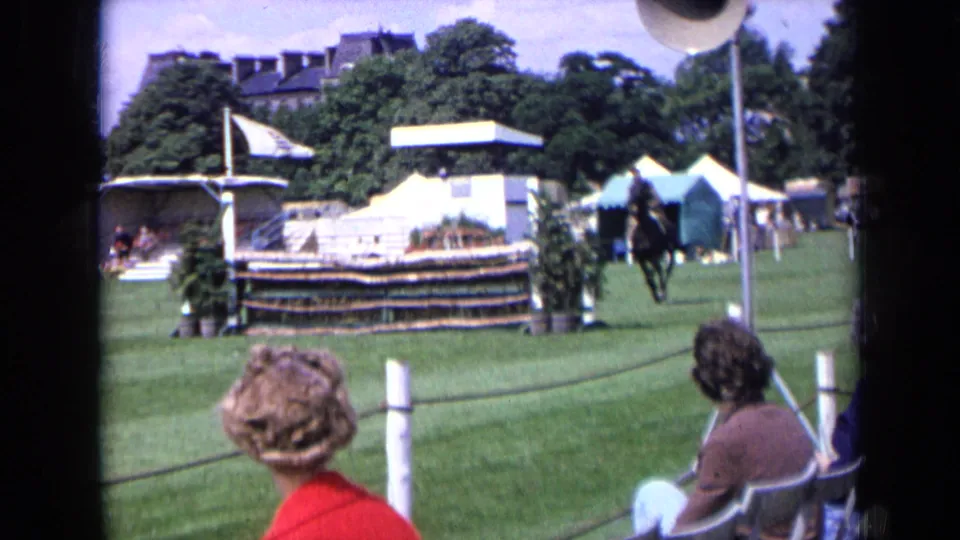 1963 England 8mm frame from the spectator side of a horse show, with seated viewers, rope barrier, white tents, and a rider near the jump area.