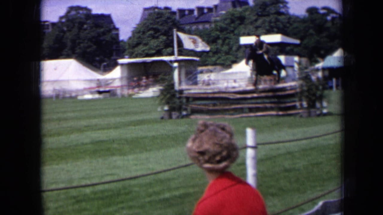 1963 England home-movie frame of a rider at a horse-jumping obstacle, with a roped spectator area, tents, trees, and field structures behind.