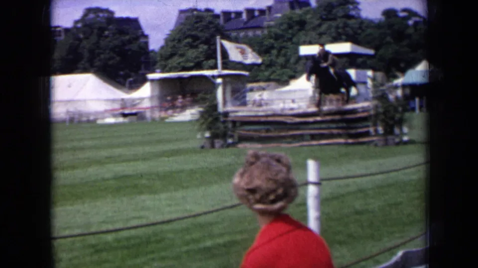 1963 England home-movie frame of a rider at a horse-jumping obstacle, with a roped spectator area, tents, trees, and field structures behind.